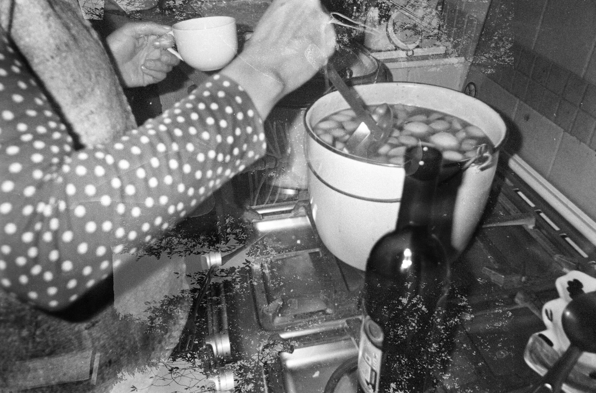 Un brazo con blusa de lunares mueve una olla llena de fruta en la estufa, con una taza en la otra mano, en blanco y negro.