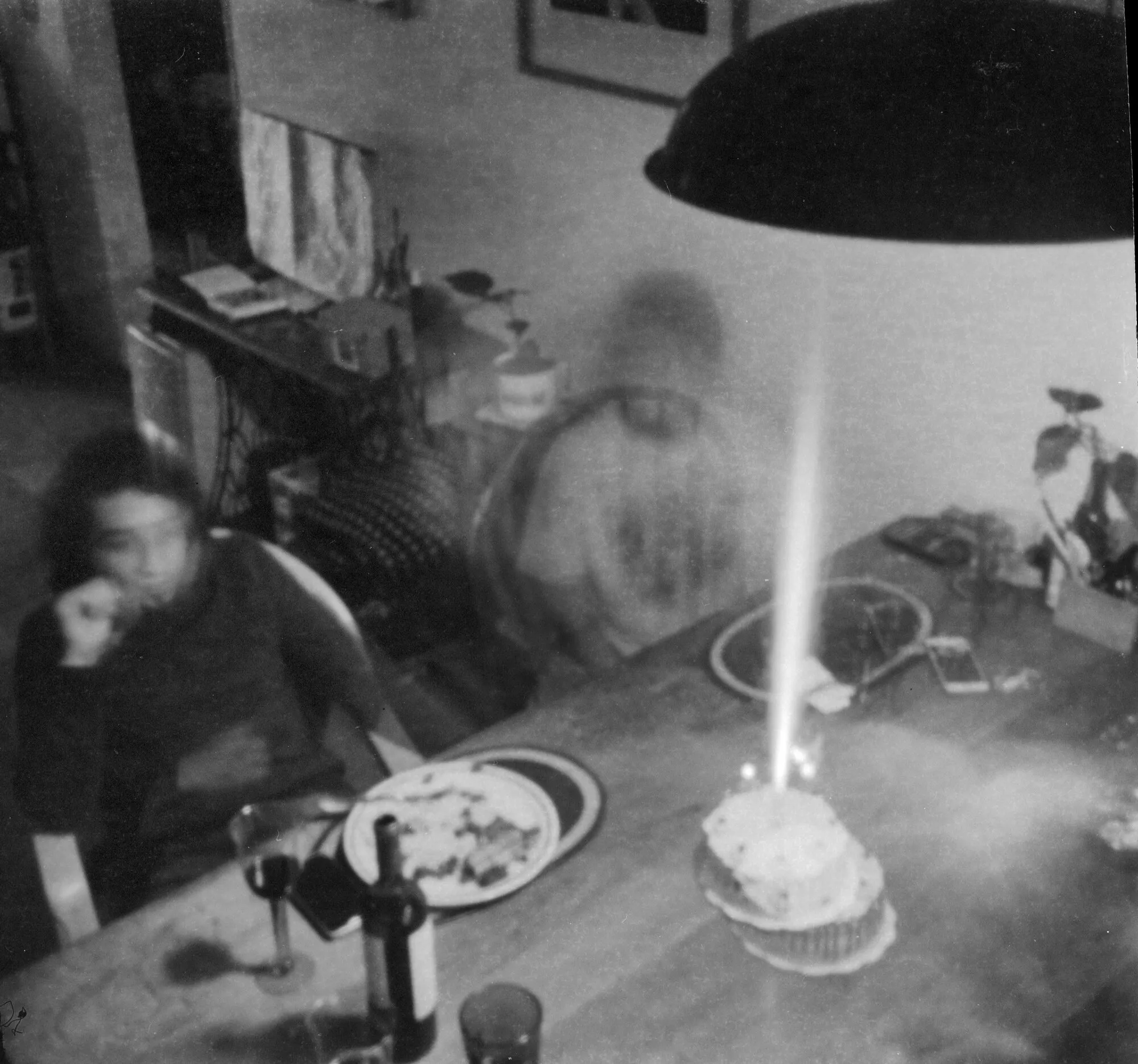 Mesa con un pastel de velas encendidas y personas movidas como fantasmas alrededor, en blanco y negro.
