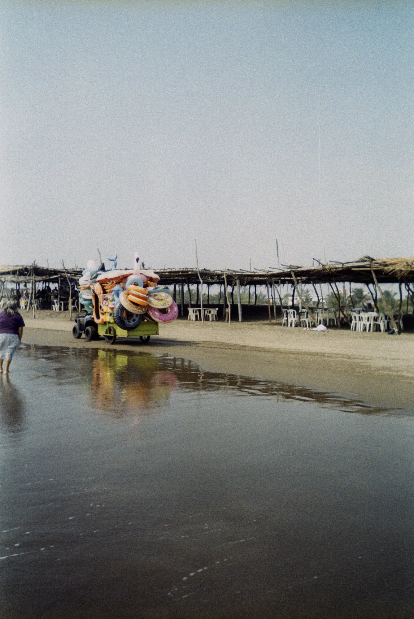 Un carrito cargado de inflables de colores en la orilla de la playa, reflejado en la arena mojada, con palapas al fondo.