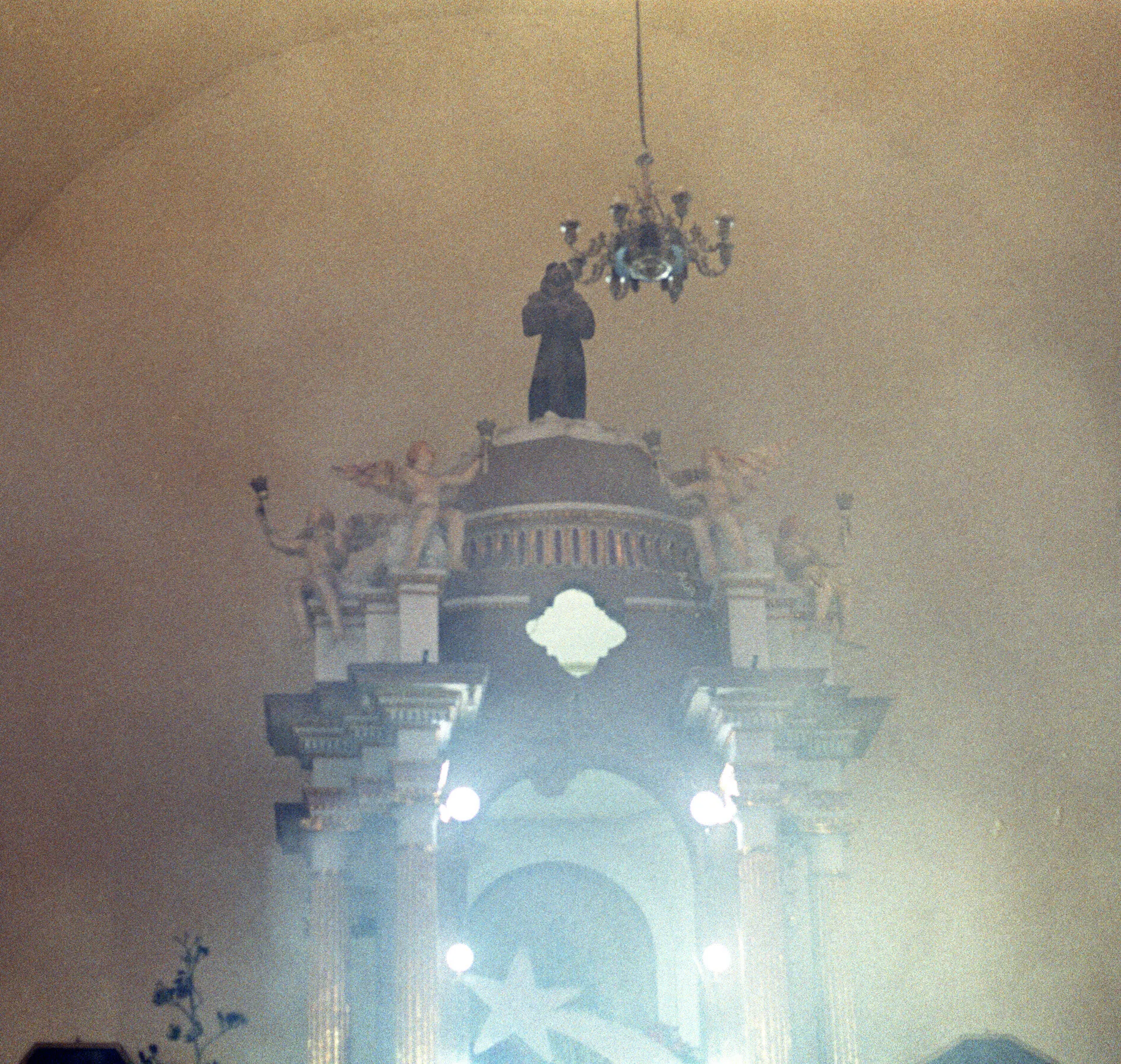 Retablo de iglesia con ángeles y un santo en lo alto, bajo un candelabro, deslavado por la luz.