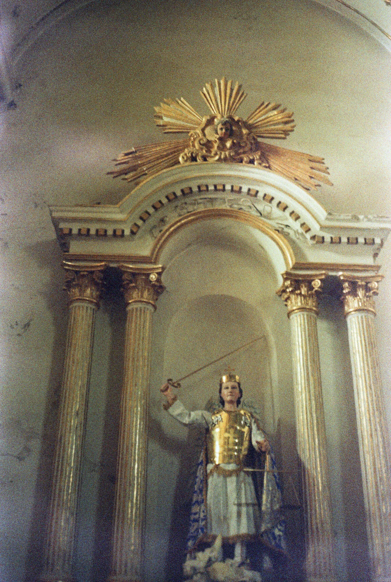 Escultura de un santo con armadura dorada y la espada en alto, en su nicho de columnas doradas.