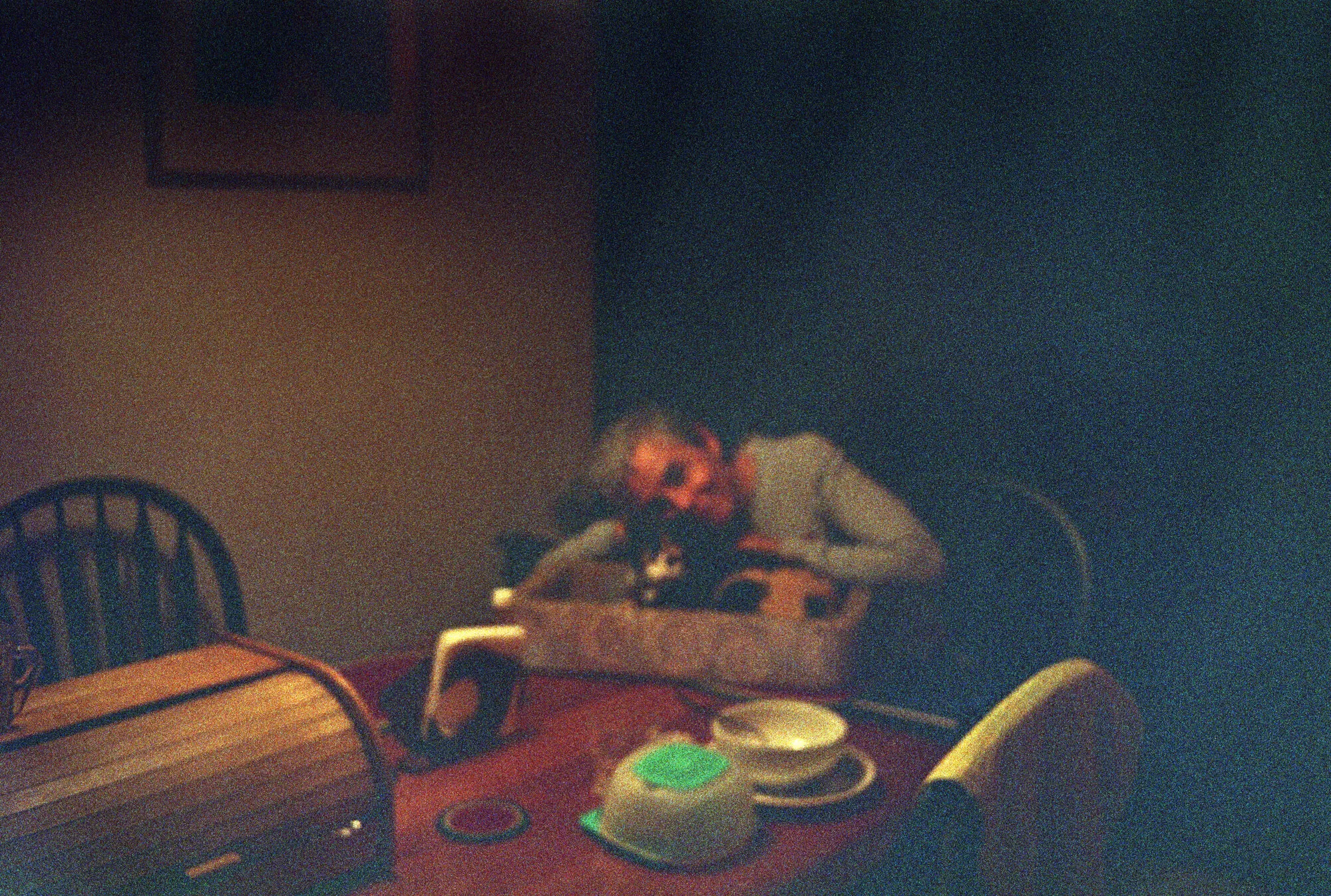 Alguien recostado sobre una mesa de mantel rojo, con tazas y una panera, en la penumbra de la noche.